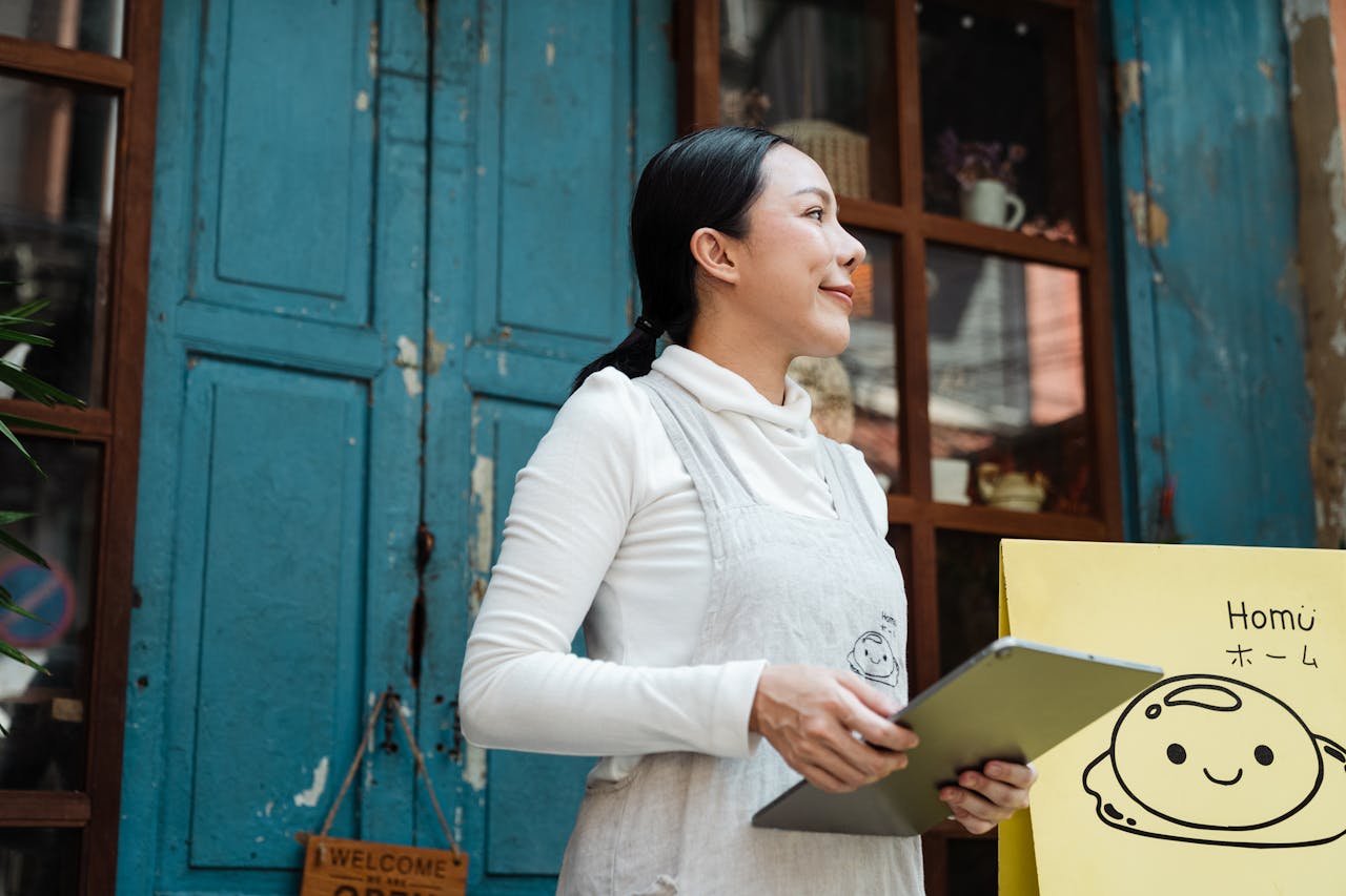 Asian woman in apron holding tablet in front of a blue door with signage.
